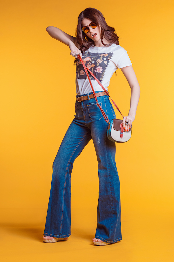 Beautiful young brunette girl wearing white t-shirt jeans flared with a small bag standing on a yellow background in the Studio.