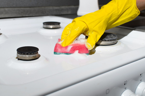 Female hand in glove cleaning the surface of gas stove with washing sponge and chemical foam. Home cleaning concept, kitchen hygiene